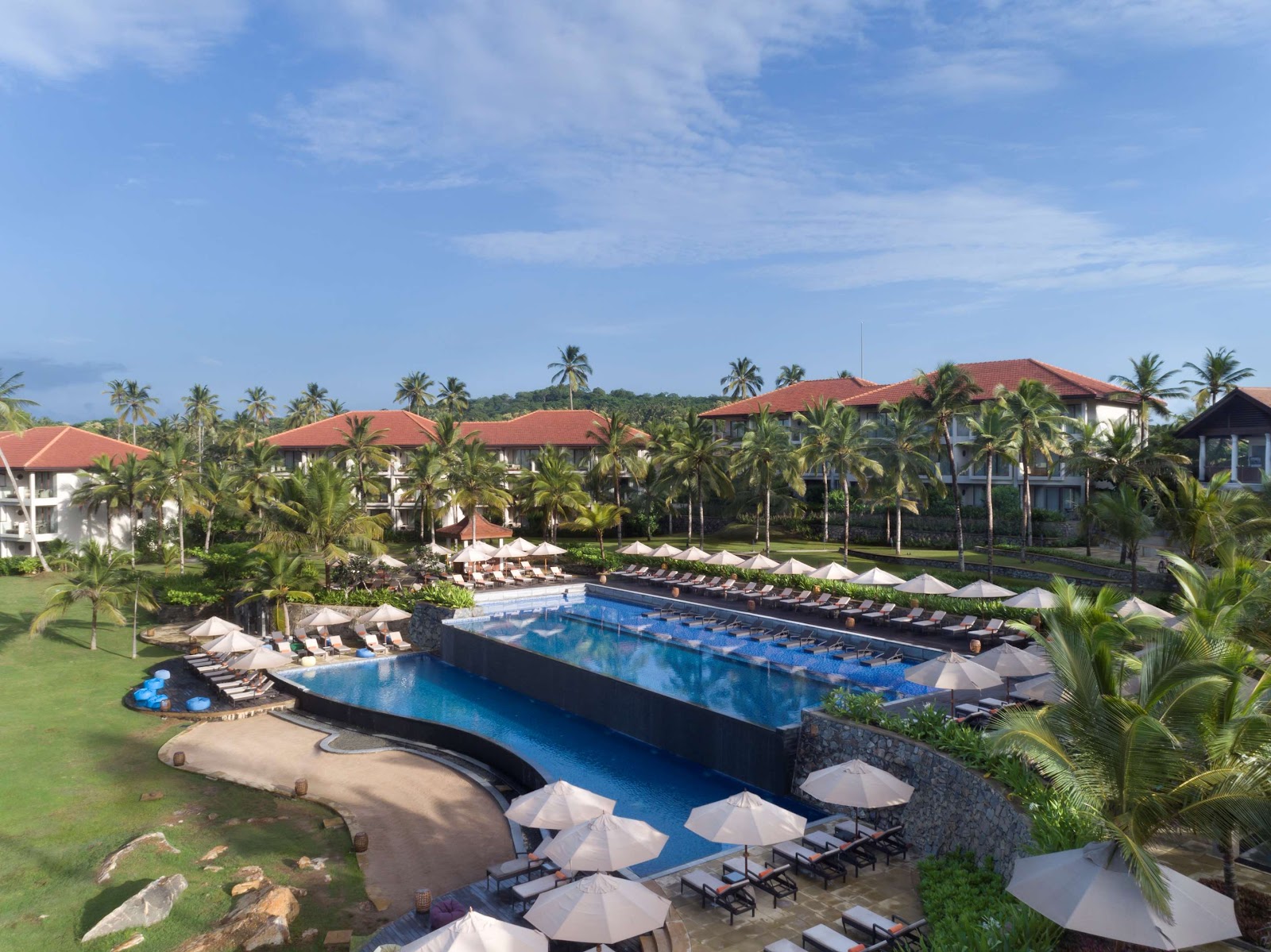 Anantara Peace Haven Tangalle Resort pool complex with red-roofed buildings, palm trees, loungers, and white umbrellas under blue sky.