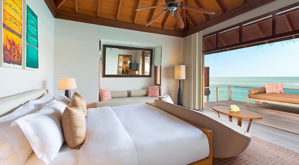 Overwater bungalow bedroom at Anantara Veli Maldives with queen bed, wooden ceiling fan, and turquoise ocean view through open deck doors.