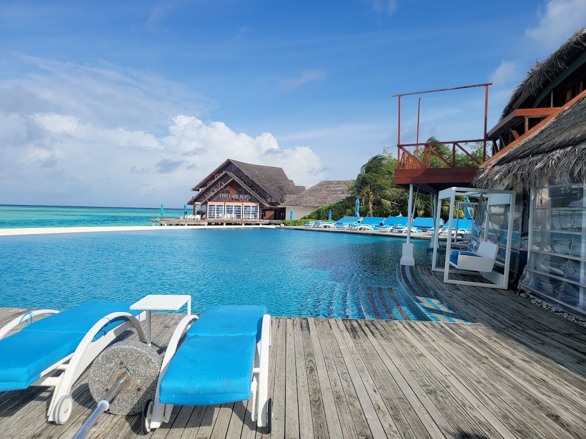 Blue loungers line the wooden deck beside turquoise lagoon water, with thatched-roof overwater bungalows and a beach visible across the bay.