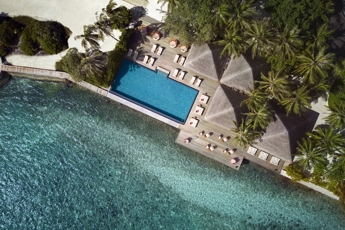 Aerial view of a resort pool deck with turquoise ocean, wooden loungers, palm trees, and guests relaxing by the water.