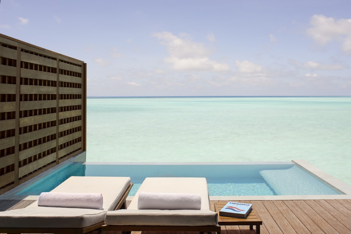 Two white loungers with rolled towels sit on a wooden deck beside a pool overlooking turquoise ocean water at Anantara Veli Maldives Resort.
