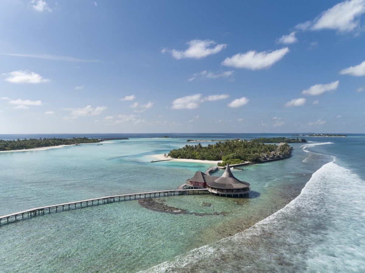Aerial view of overwater bungalows with thatched roofs connected by boardwalk to a tropical island lagoon ringed by palm trees.