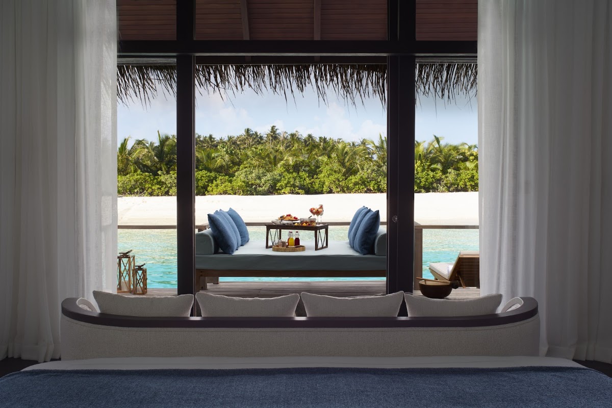 View from an overwater villa interior through open doors to a furnished deck, turquoise lagoon, and tropical island vegetation at Anantara Veli Maldives Resort.