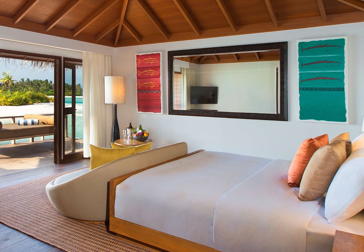 Resort bedroom with wood ceiling, white bed with orange pillows, curved chair, and open doors revealing ocean and palm trees beyond.