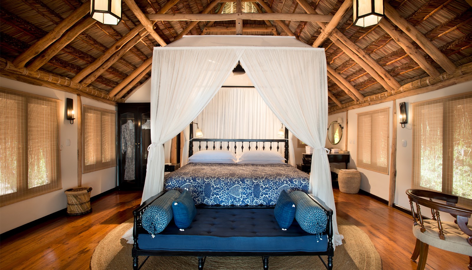 Spacious resort bedroom with wooden beams, canopied bed with white curtains, blue seating bench, and glass doors opening to natural light.