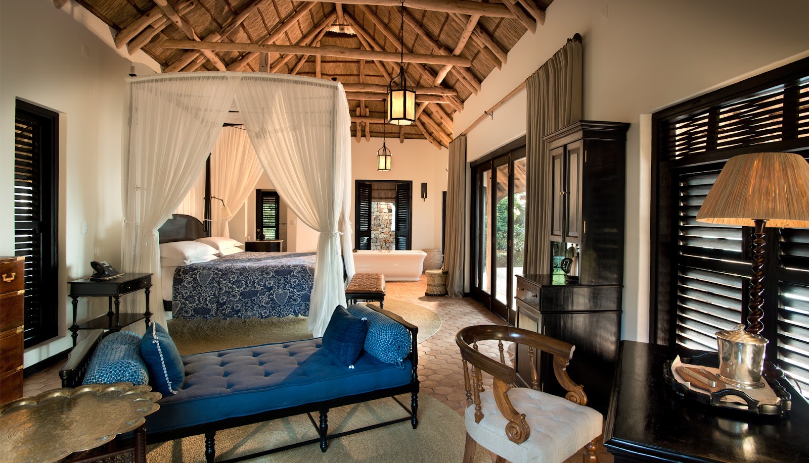 Spacious resort suite with wooden beam ceiling, four-poster canopy bed with white curtains, blue upholstered bench, black louvered doors, and warm lighting.