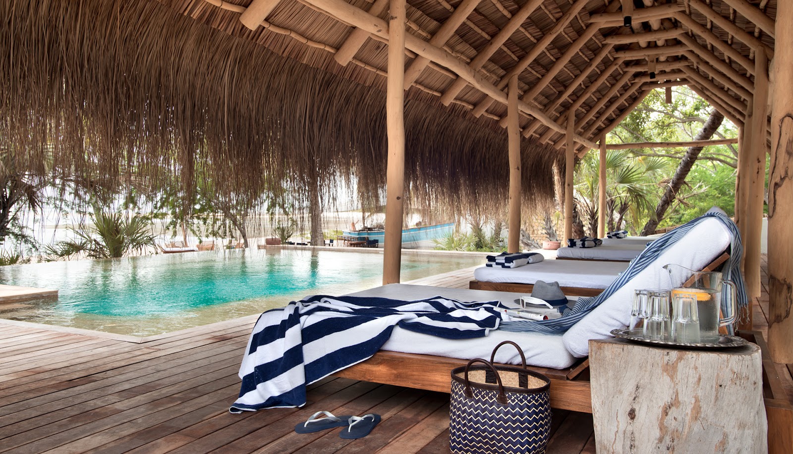 Lounge bed with striped navy and white blanket under thatched pavilion overlooking turquoise lagoon at andBeyond Benguerra Island.