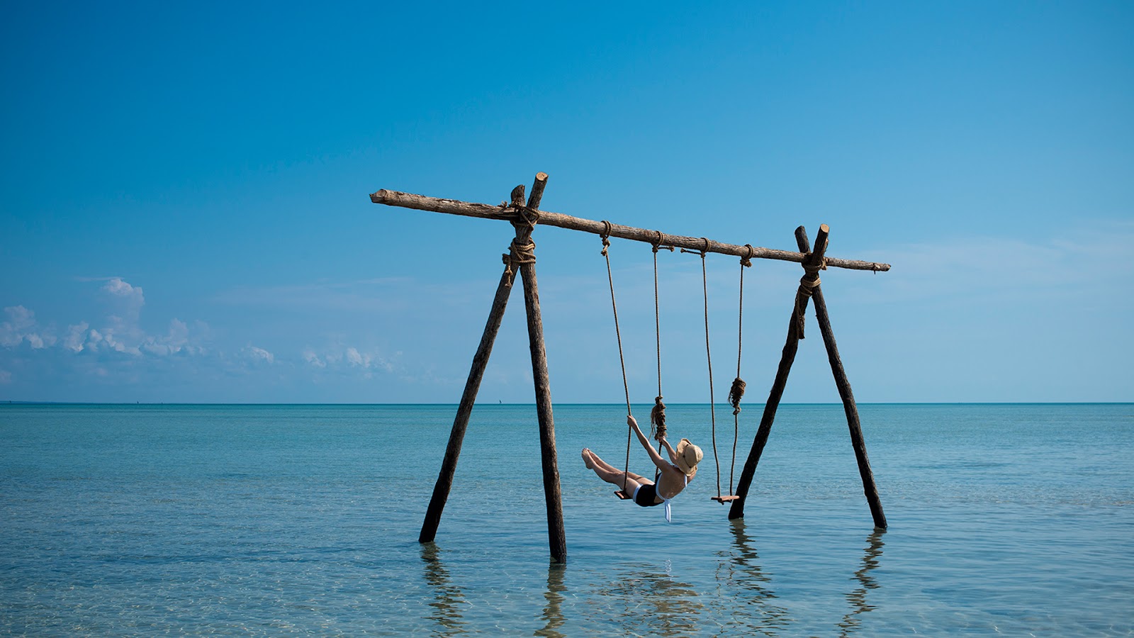 A guest swings on a wooden rope swing suspended over clear turquoise ocean water under blue sky at andBeyond Benguerra Island.
