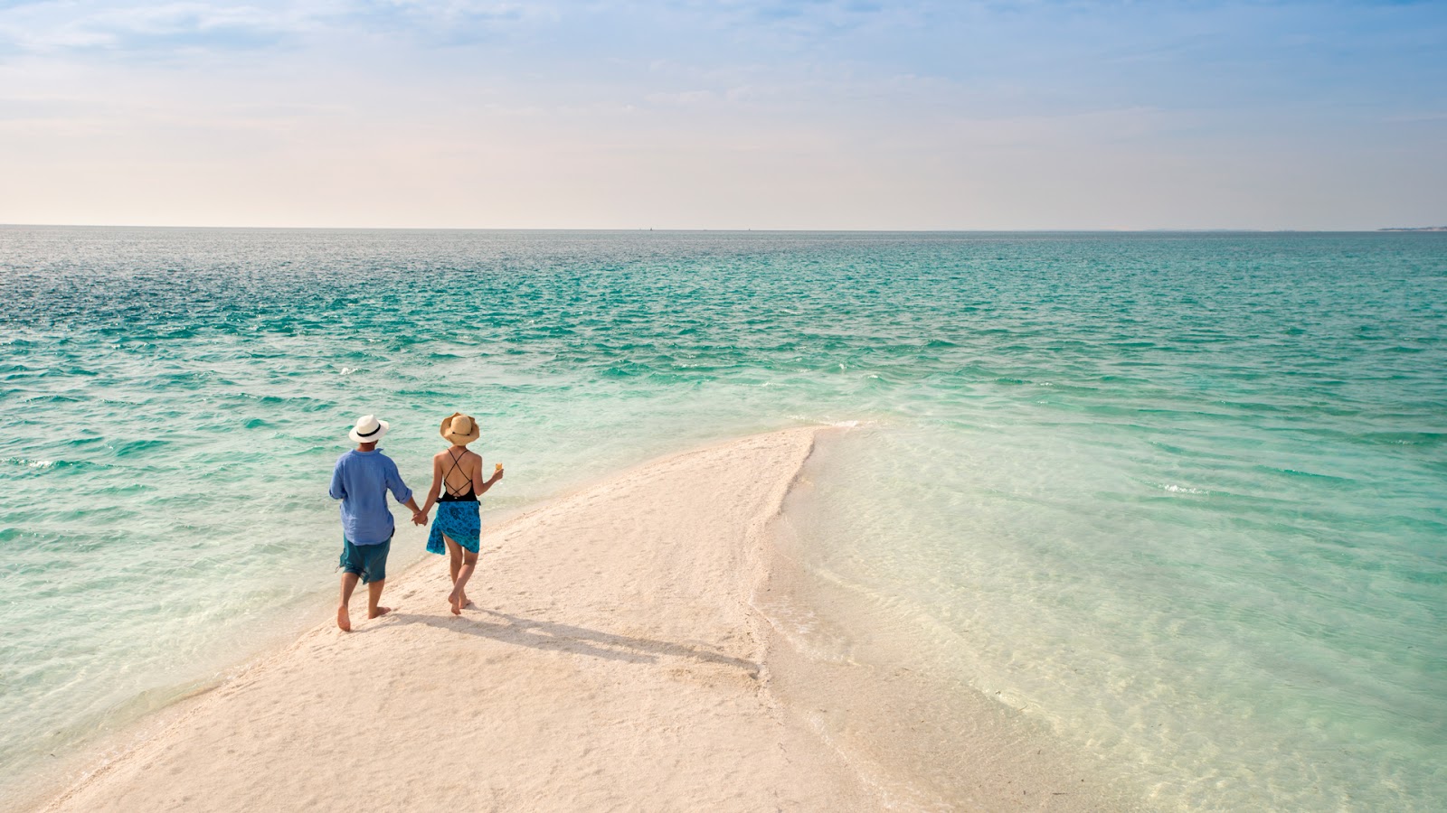 Two guests walk hand in hand on a sandbar in shallow turquoise water at andBeyond Benguerra Island.