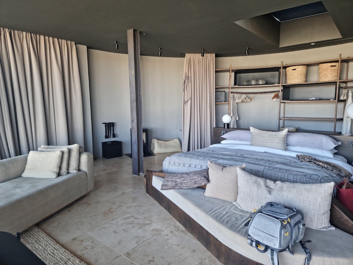 Modern resort suite with concrete floors, four-poster bed, open shelving, cream curtains, and a seating area with neutral furnishings.