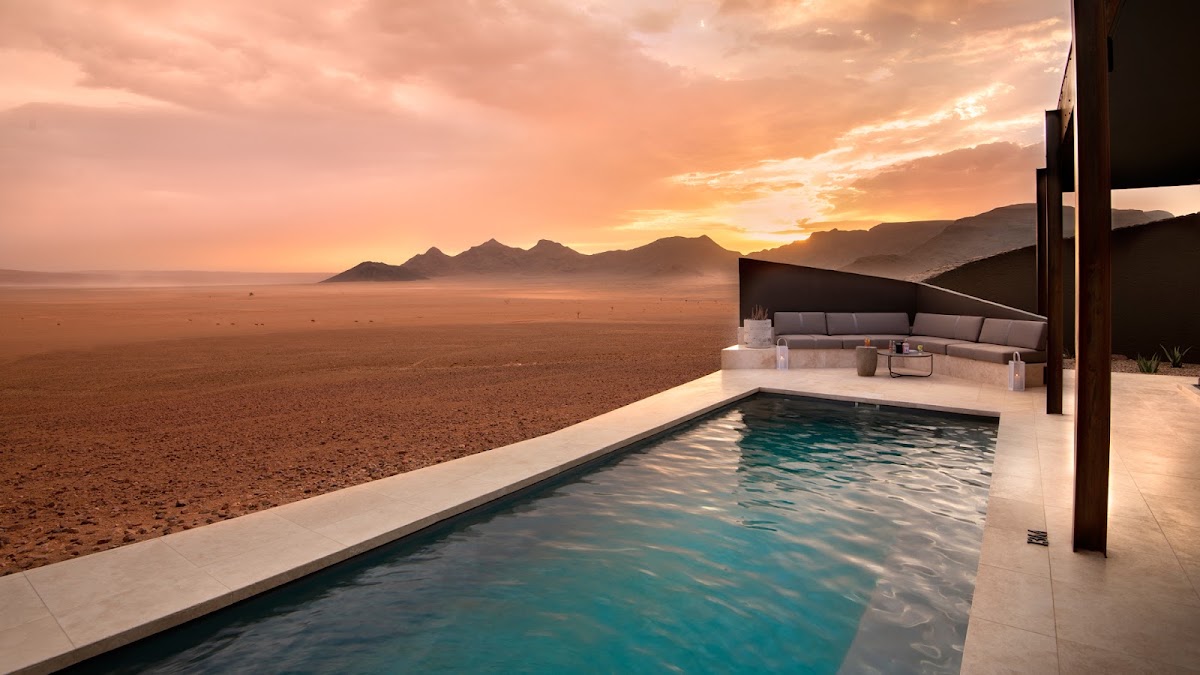 Pool overlooking vast desert at sunset with distant mountains and ocean horizon at &Beyond Sossusvlei Desert Lodge.