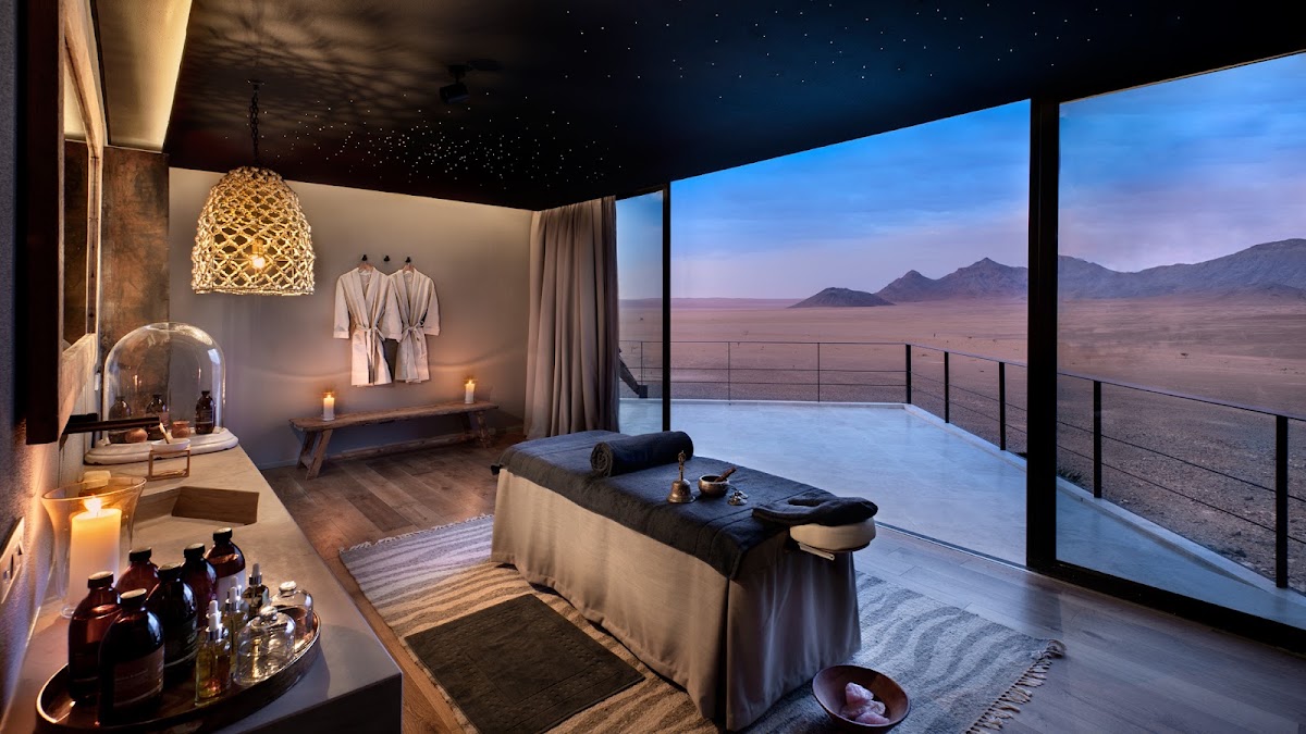 Spa treatment room at &Beyond Sossusvlei Desert Lodge with massage bed, floor-to-ceiling windows framing desert landscape and distant mountains at dusk.