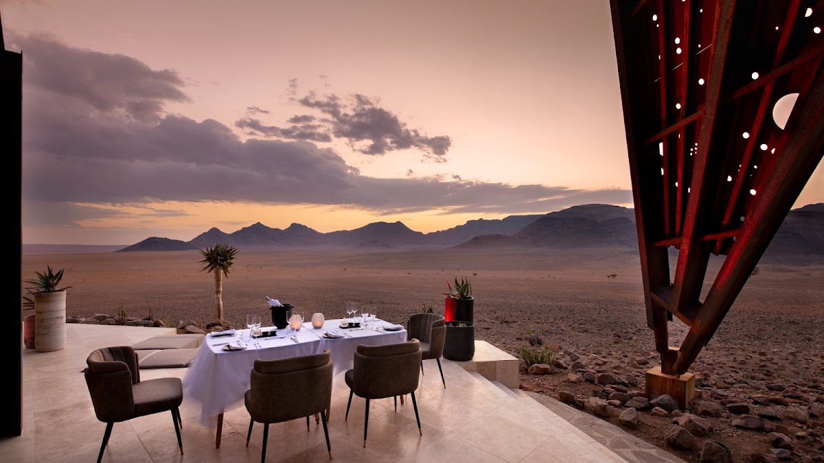 Outdoor dining table set for two on a stone terrace overlooking desert dunes and mountains at sunset, with a red metal pergola overhead.