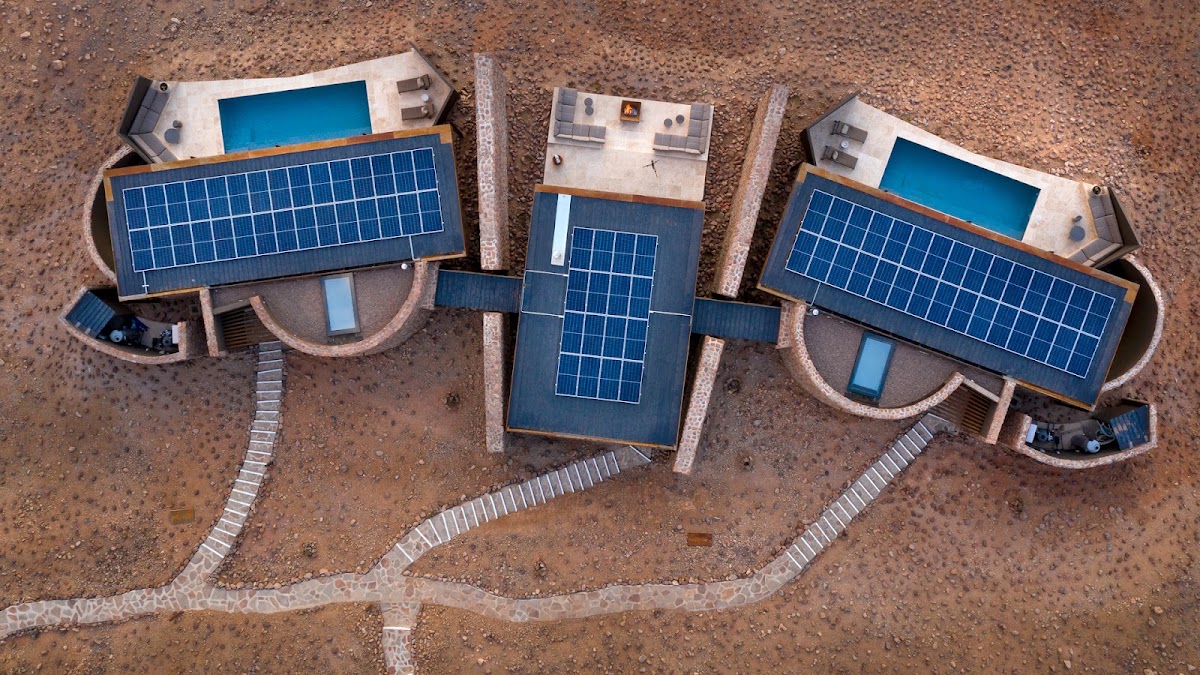 Aerial view of three curved guest suites with solar panels on their roofs and blue plunge pools, arranged in the red desert sand at &Beyond Sossusvlei Desert Lodge.