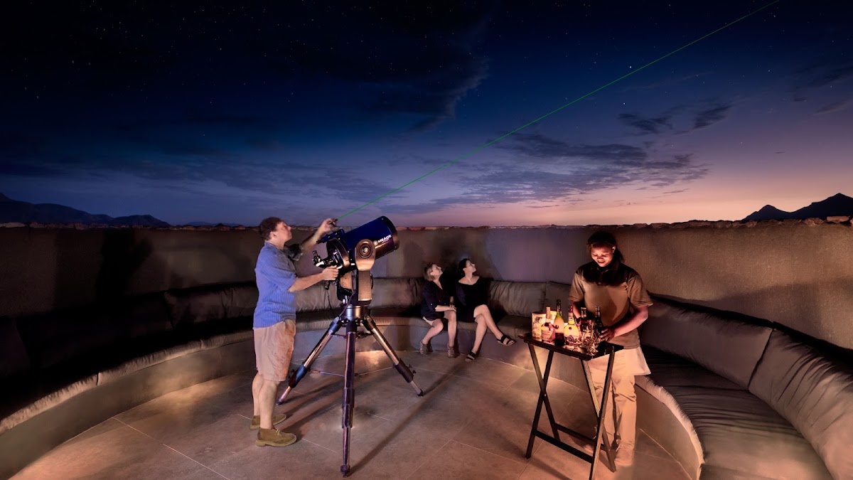 Guests stargazing on a rooftop terrace at dusk, with one person viewing through a telescope while others relax on seating nearby.