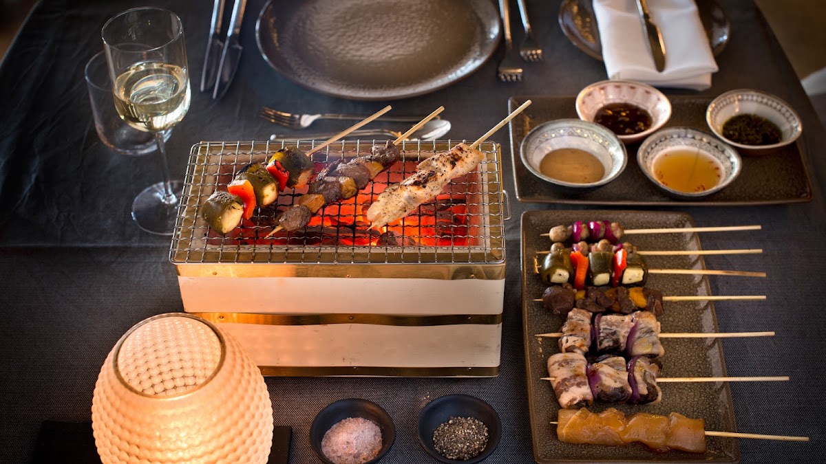Tabletop grill station with meat and vegetable skewers cooking over flames, surrounded by condiment bowls, raw skewers, and place settings at &Beyond Sossusvlei Desert Lodge.