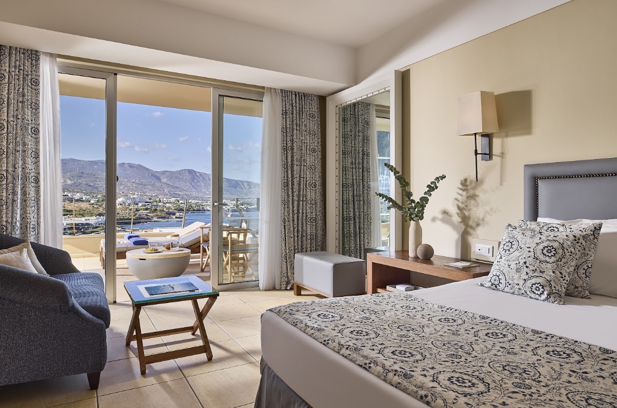 Resort bedroom with patterned bedding, seating area, and glass doors opening to a balcony overlooking a coastal bay and mountains.