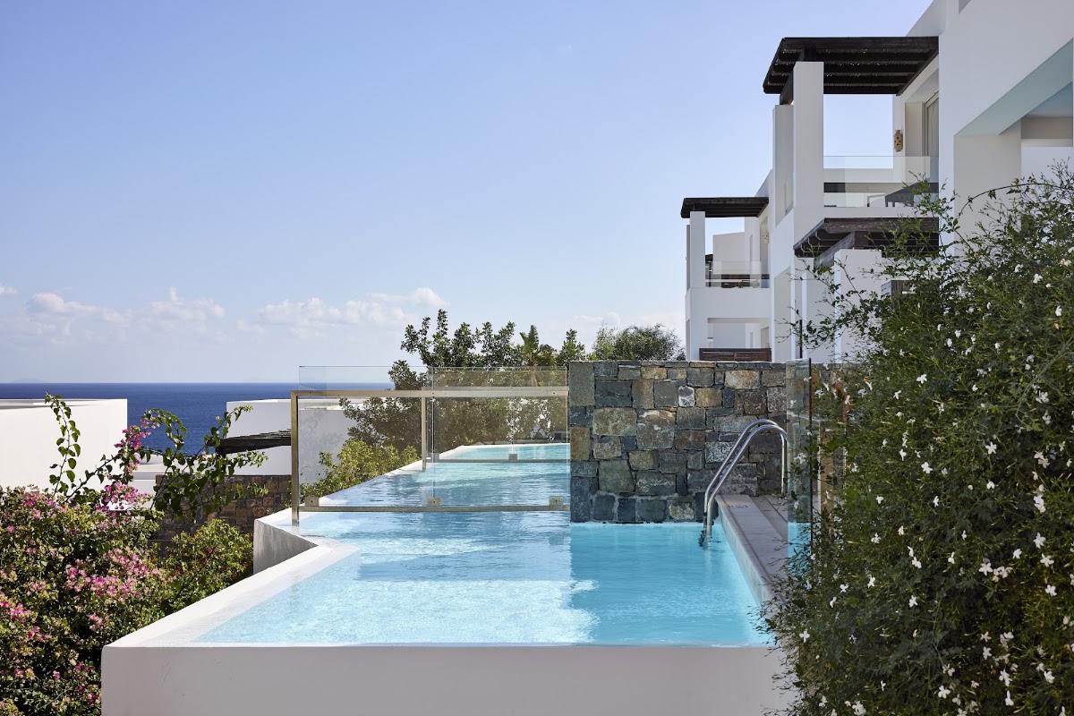 Infinity pool with turquoise water overlooking the sea at Aquila Elounda Village, flanked by white resort buildings and flowering plants.