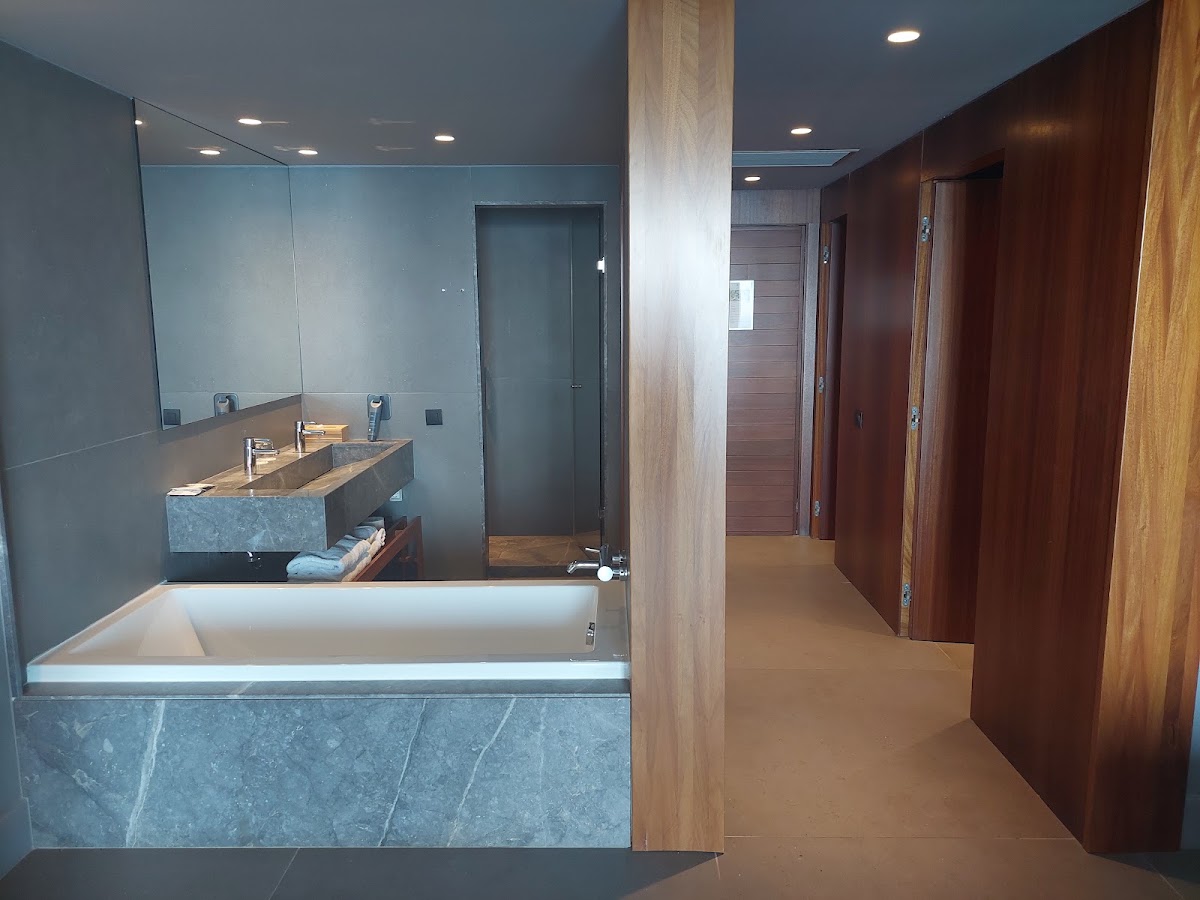 Modern bathroom with stone bathtub, dual sink vanity, glass shower enclosure, and wooden doors leading to bedroom areas.