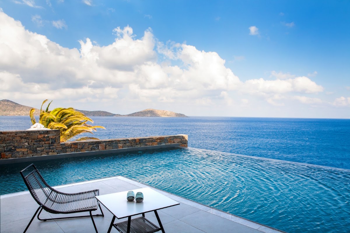 A black woven lounge chair and white table with slippers sit on a terrace overlooking turquoise water and distant islands under a blue sky with white clouds.