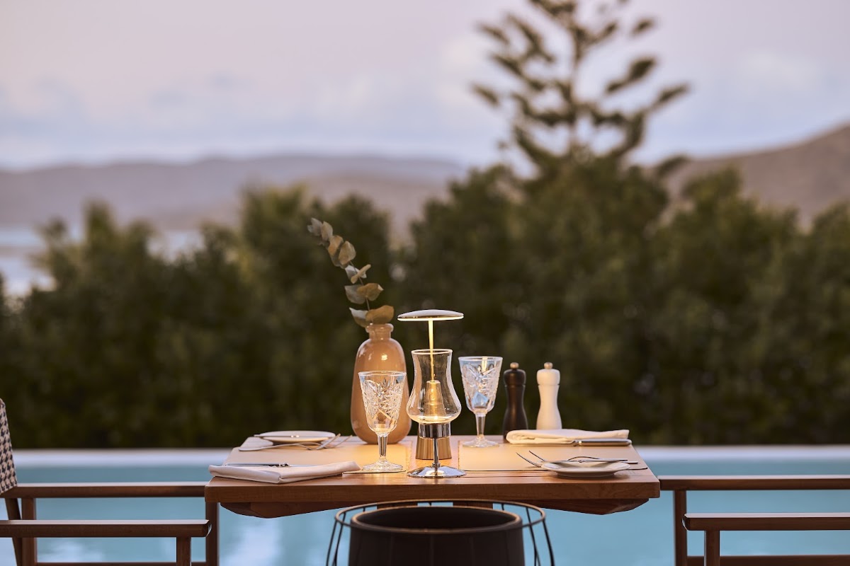 Dining table set with glasses and place settings overlooking a pool and forested hills at Aquila Elounda Village.
