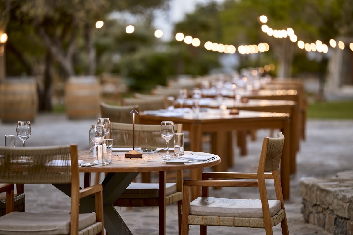Outdoor dining area at Aquila Elounda Village with wooden tables, place settings, and string lights above trees at dusk.