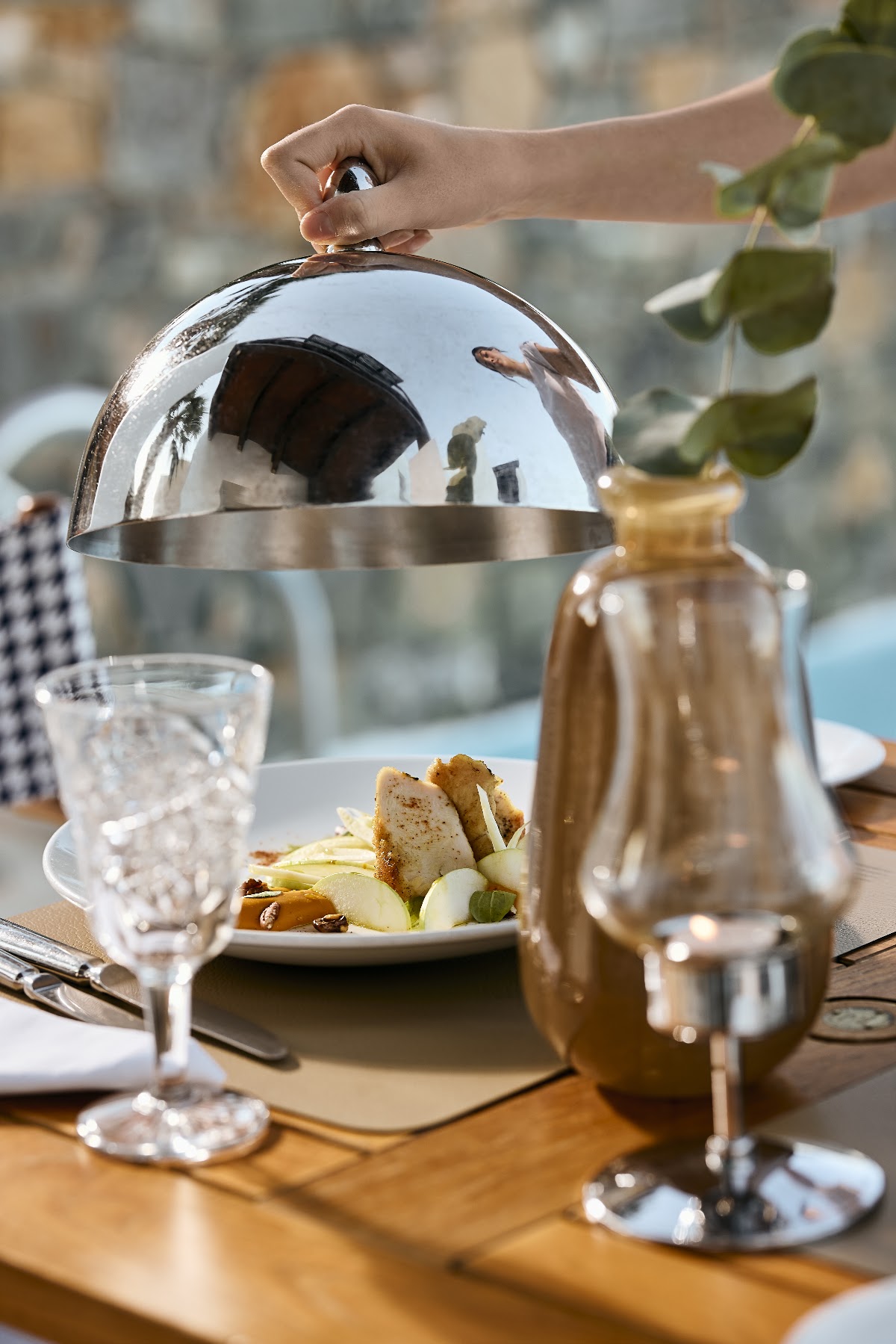 A server's hand lifts a silver cloche from a plated dish of roasted vegetables at an outdoor dining table overlooking resort buildings.