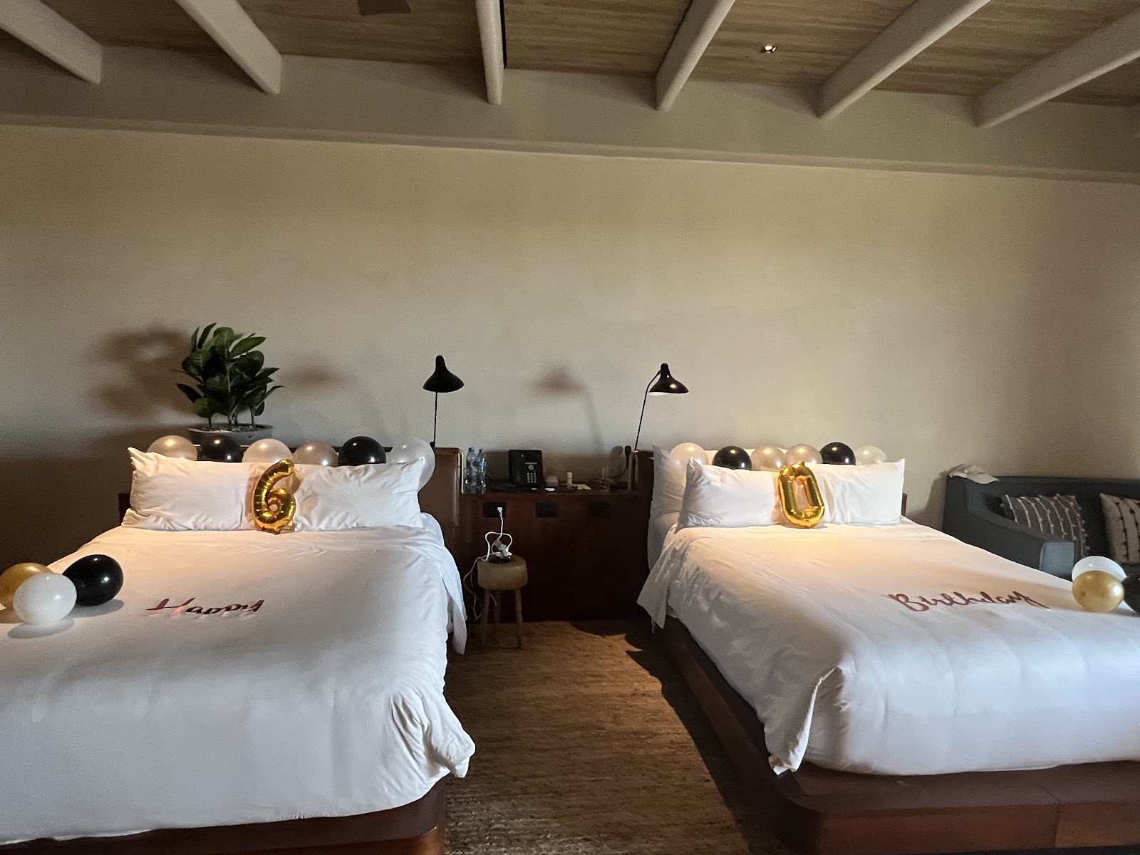 Two queen beds with white linens and gold balloon decorations in a modern resort room with wooden beams and black task lamps.