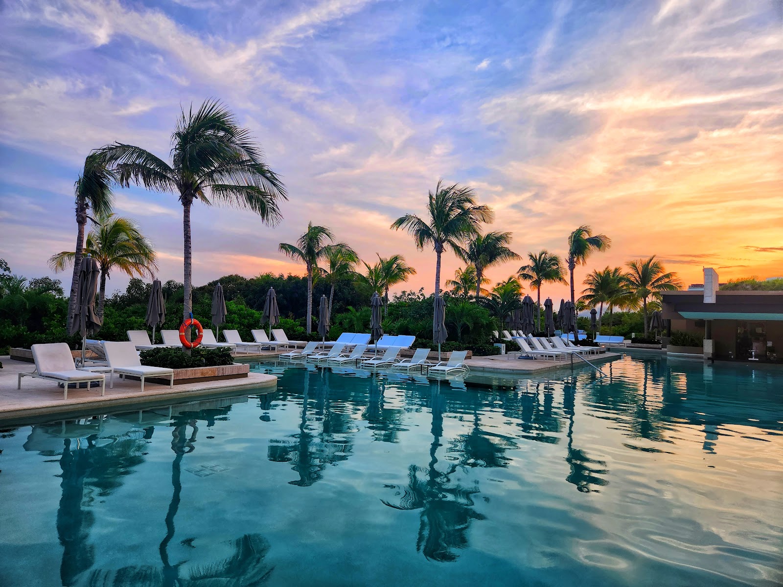 Resort pool at sunset with lounge chairs, palm trees, and closed umbrellas along the deck as the sky turns orange and pink.