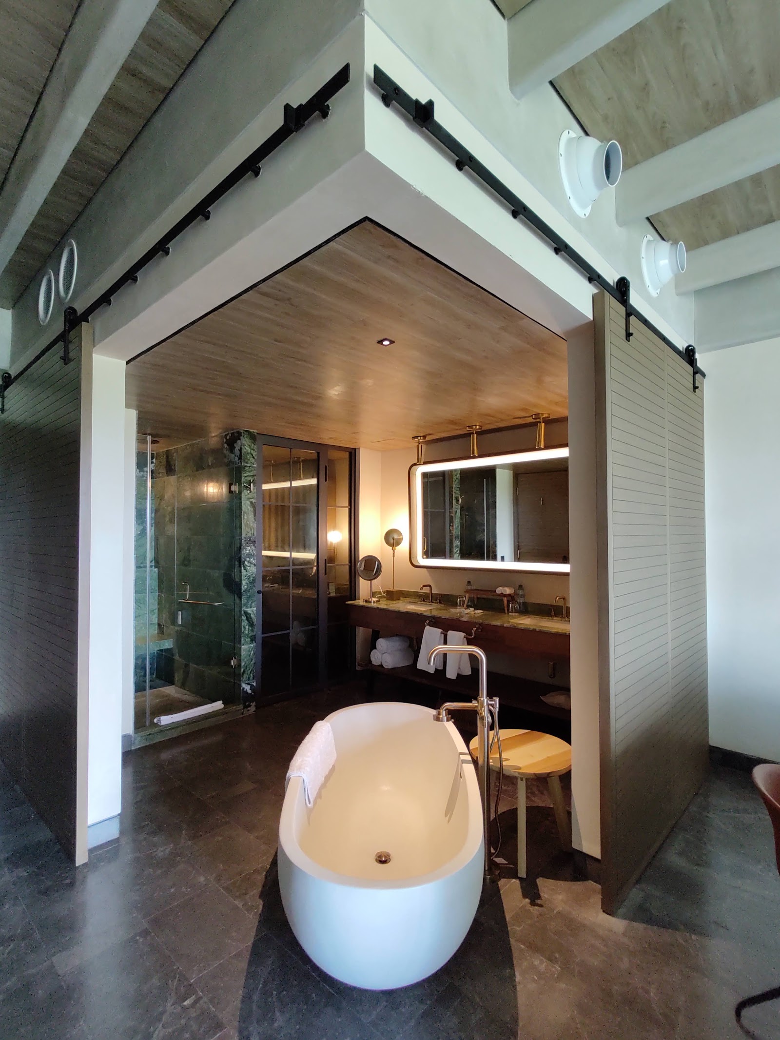Modern resort bathroom with freestanding white tub, glass shower enclosure, illuminated mirror, and wooden vanity under exposed ceiling beams.