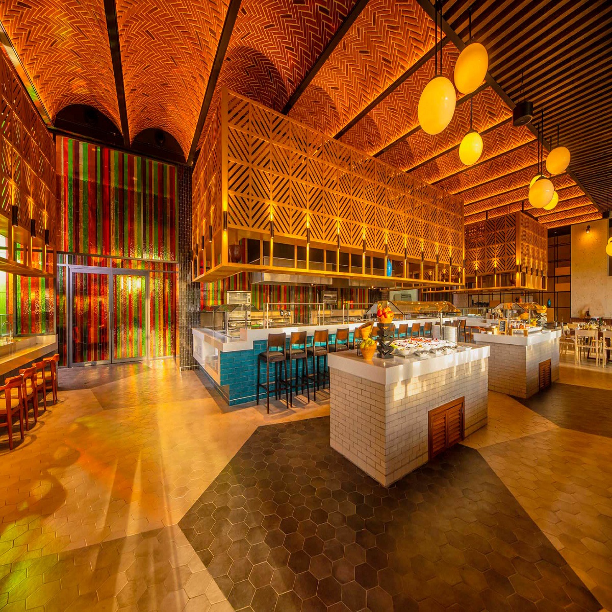 Open-plan resort kitchen with orange patterned ceilings, white brick islands, teal bar seating, and yellow pendant lights.