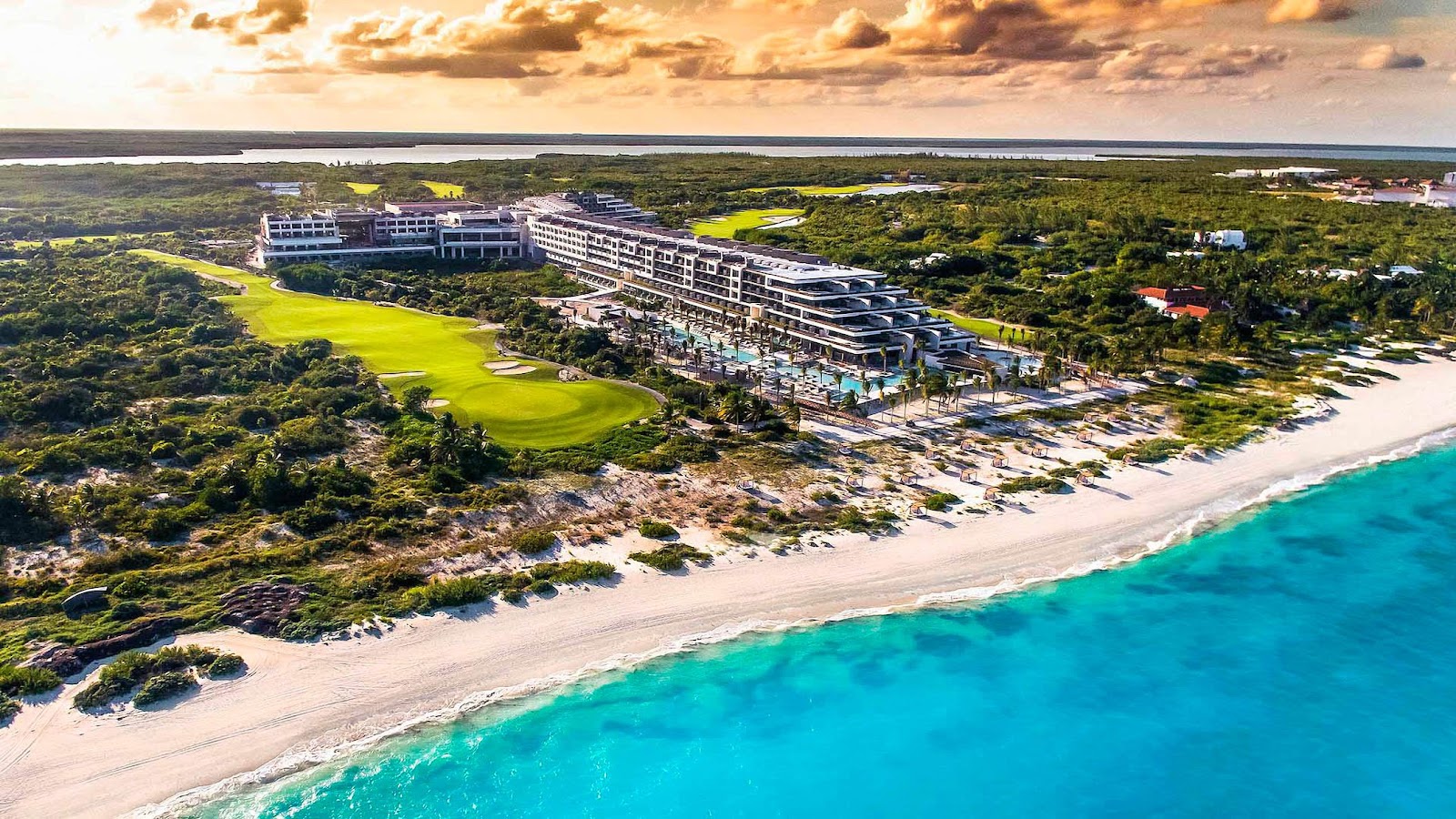 Aerial view of ATELIER Playa Mujeres resort stretching along a white sand beach with turquoise water, golf course, and dense jungle inland at sunset.