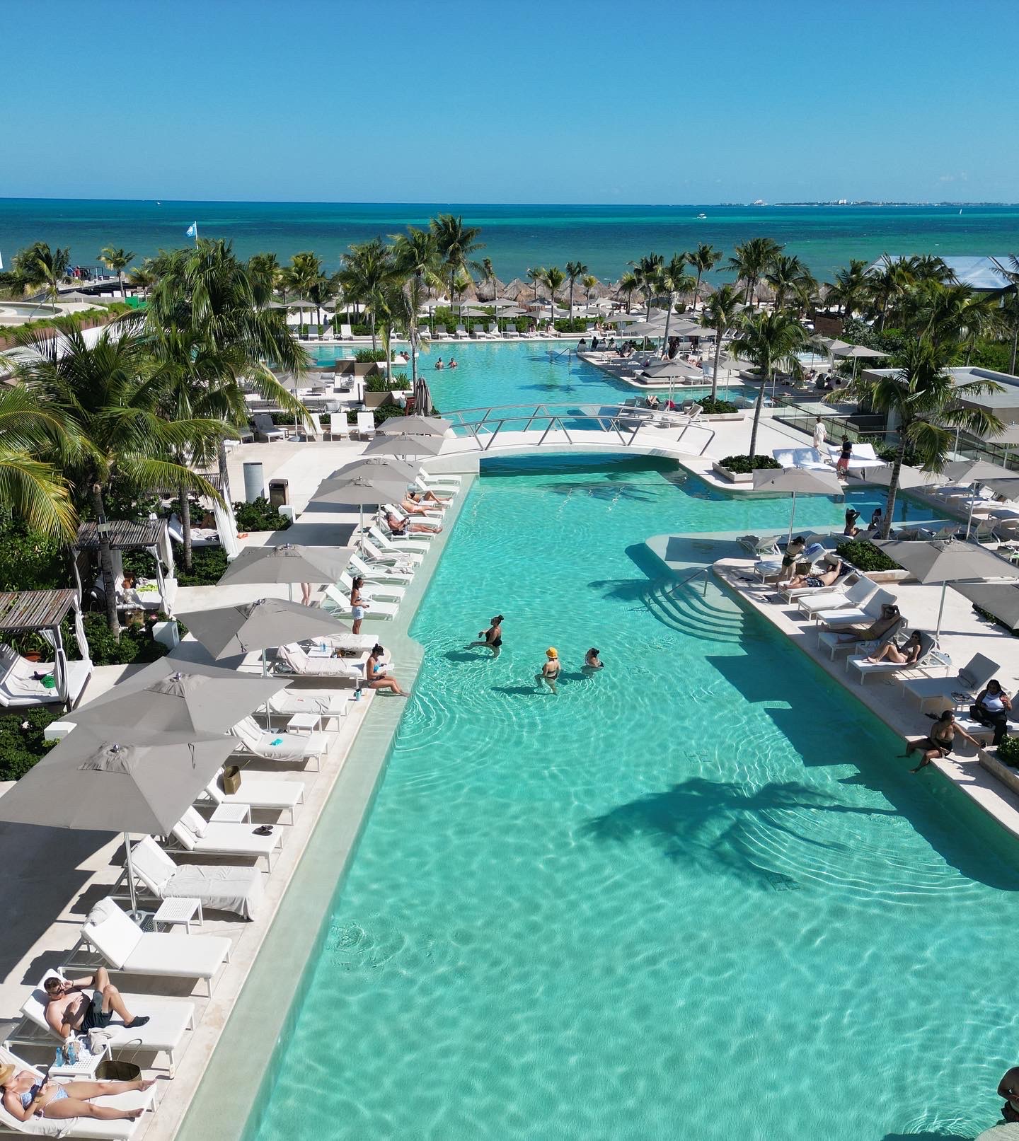 Turquoise pools lined with white lounge chairs and gray umbrellas at ATELIER Playa Mujeres, with beach and ocean beyond.