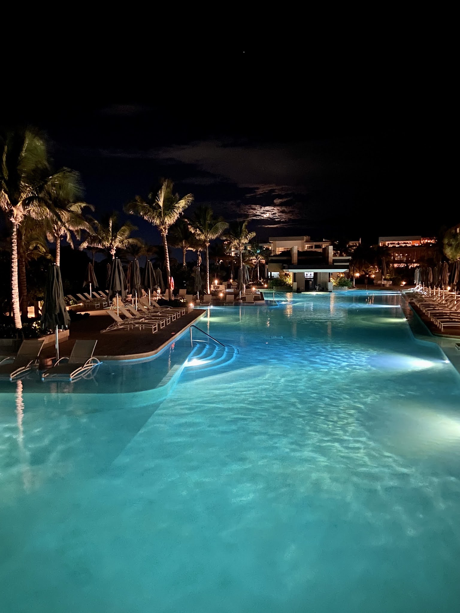 Illuminated resort pool at night with turquoise water, lounge chairs, umbrellas, palm trees, and moonlit sky above the main building.