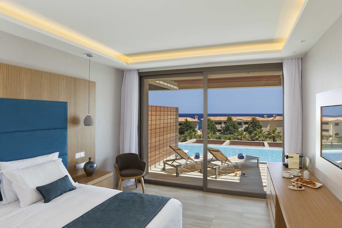 Resort bedroom with ocean view through sliding glass doors opening to a terrace with lounge chairs and pool beyond.
