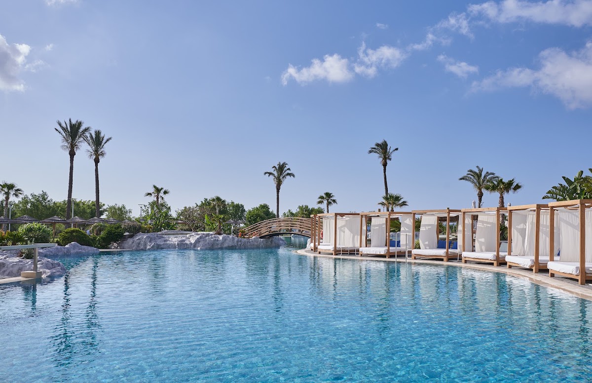Large resort pool with turquoise water, white cabanas lining the right side, palm trees throughout, and a curved wooden bridge crossing the pool under blue sky.