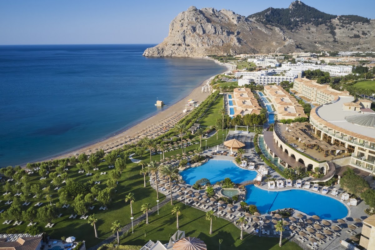 Atlantica Imperial Resort and Spa stretches across manicured grounds with two blue pools, beach loungers, and palm trees, overlooking a sandy beach and dramatic rocky headland.