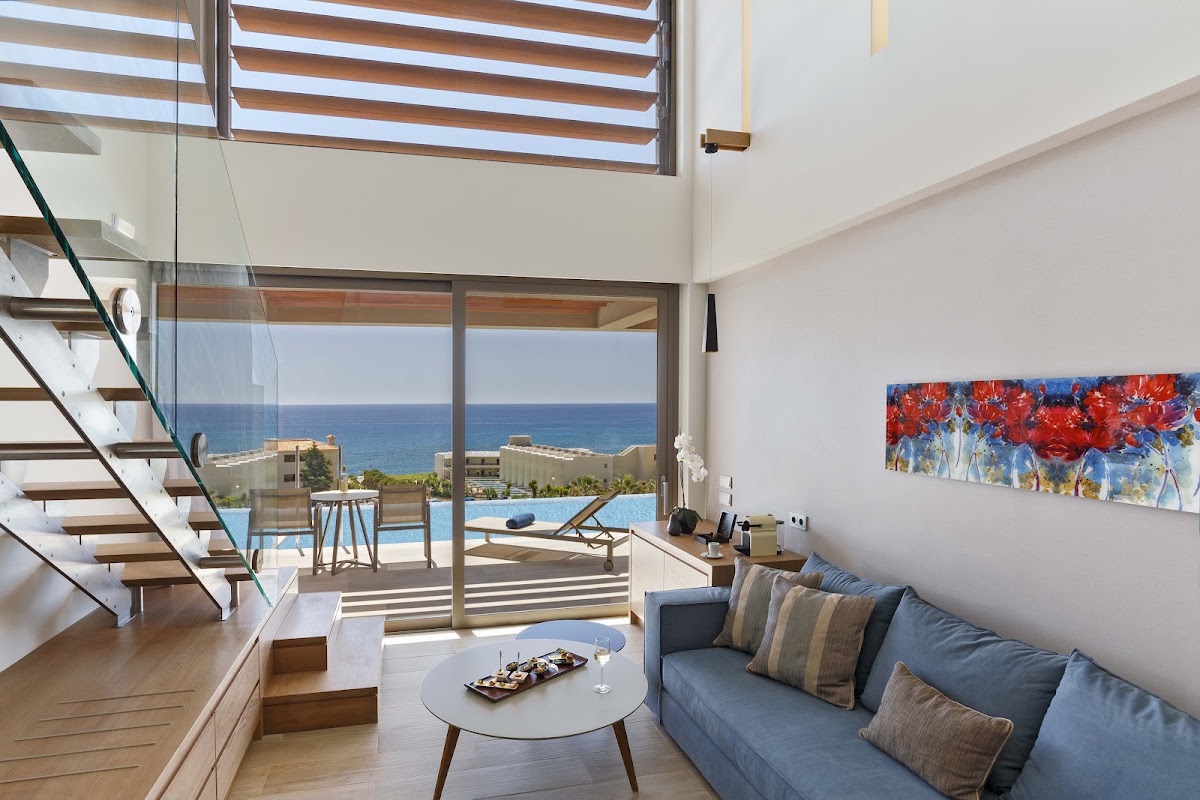 Modern resort suite with blue sofa, spiral staircase, and floor-to-ceiling windows overlooking pool and Mediterranean Sea at Atlantica Imperial Resort and Spa.