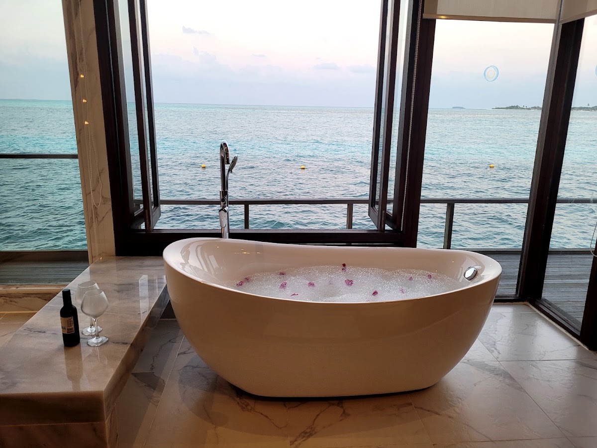 White freestanding bathtub with flower petals inside, positioned in front of floor-to-ceiling windows overlooking turquoise ocean at Atmosphere Kanifushi Maldives.