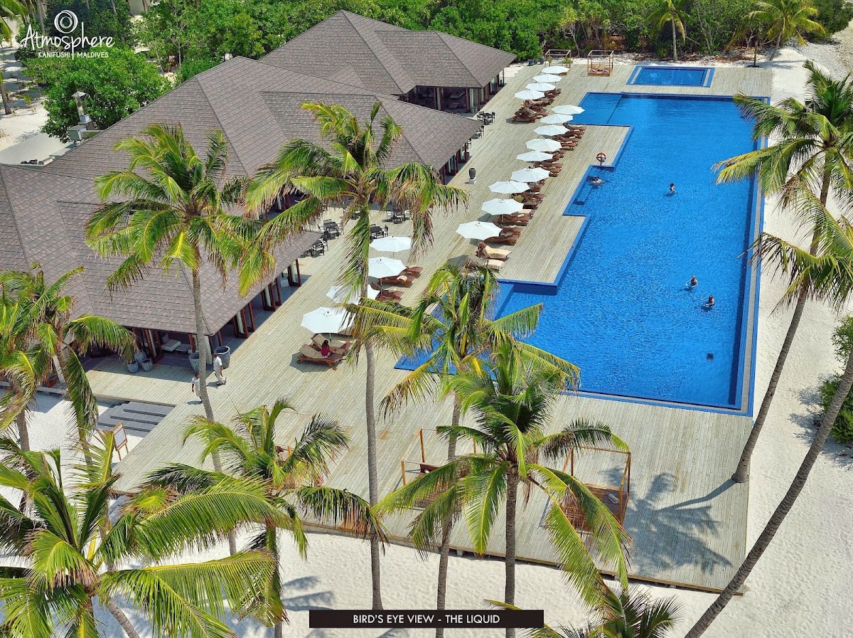 Atmosphere Kanifushi Maldives aerial view showing a large blue swimming pool with loungers, a gray-roofed main building, and palm trees surrounding the wooden deck.