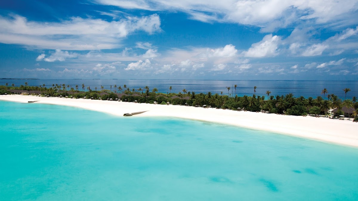 Turquoise lagoon and white sand beach lined with palm trees and resort buildings under blue sky with white clouds.