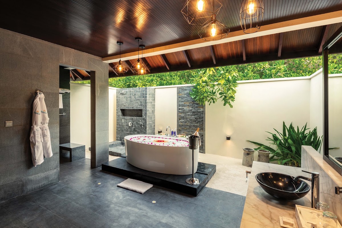 Luxury bathroom with soaking tub filled with rose petals, rainfall shower, black vessel sink, and open-air courtyard with tropical plants at Atmosphere Kanifushi Maldives.