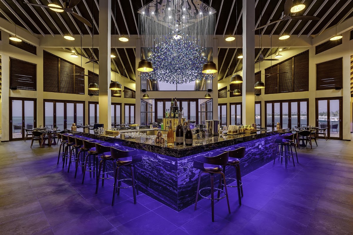 Upscale bar lounge with purple-lit countertop, crystal chandelier, floor-to-ceiling ocean views, and modern seating at Atmosphere Kanifushi Maldives.