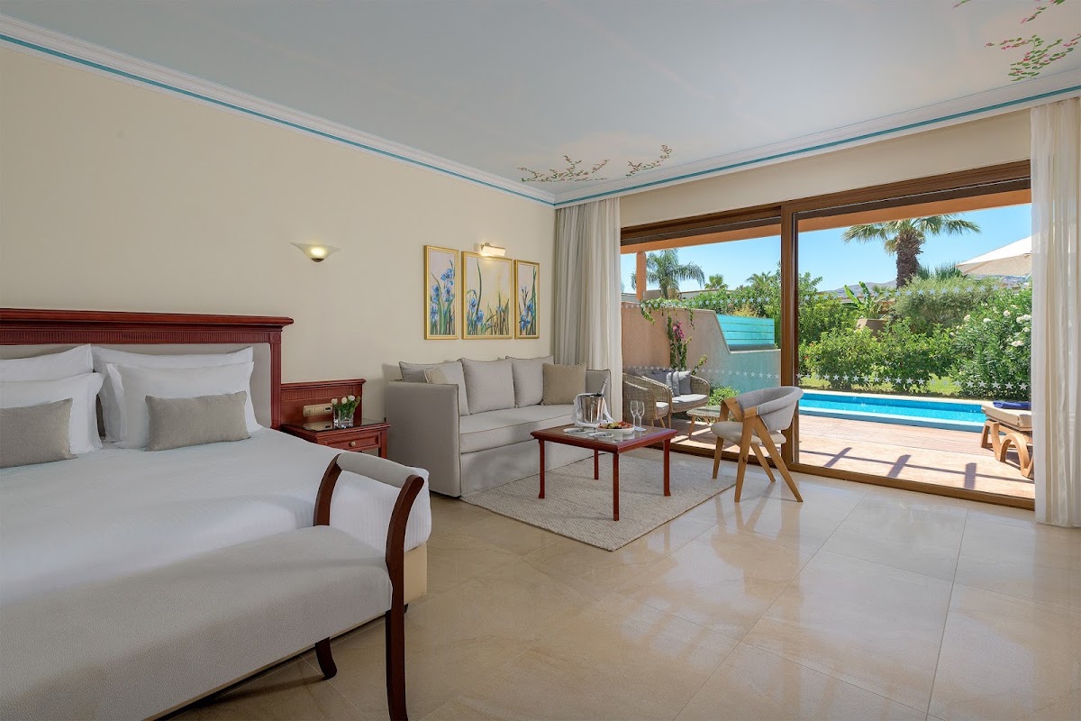 Resort suite with bed, seating area, and terrace doors opening to pool and garden views at Atrium Palace Thalasso Spa Resort & Villas.