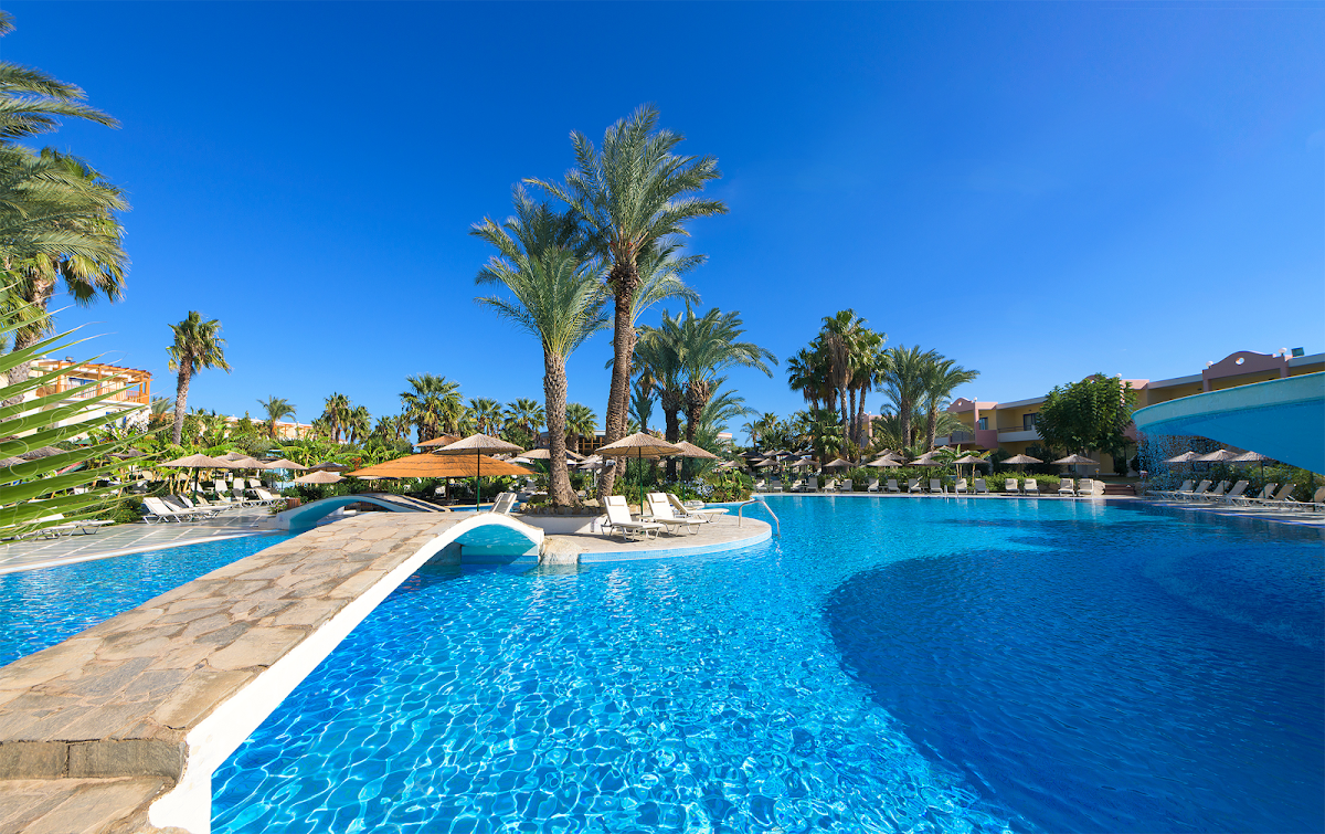 Large resort pool with curved white bridge, palm trees, loungers with tan umbrellas, and hotel buildings under clear blue sky.