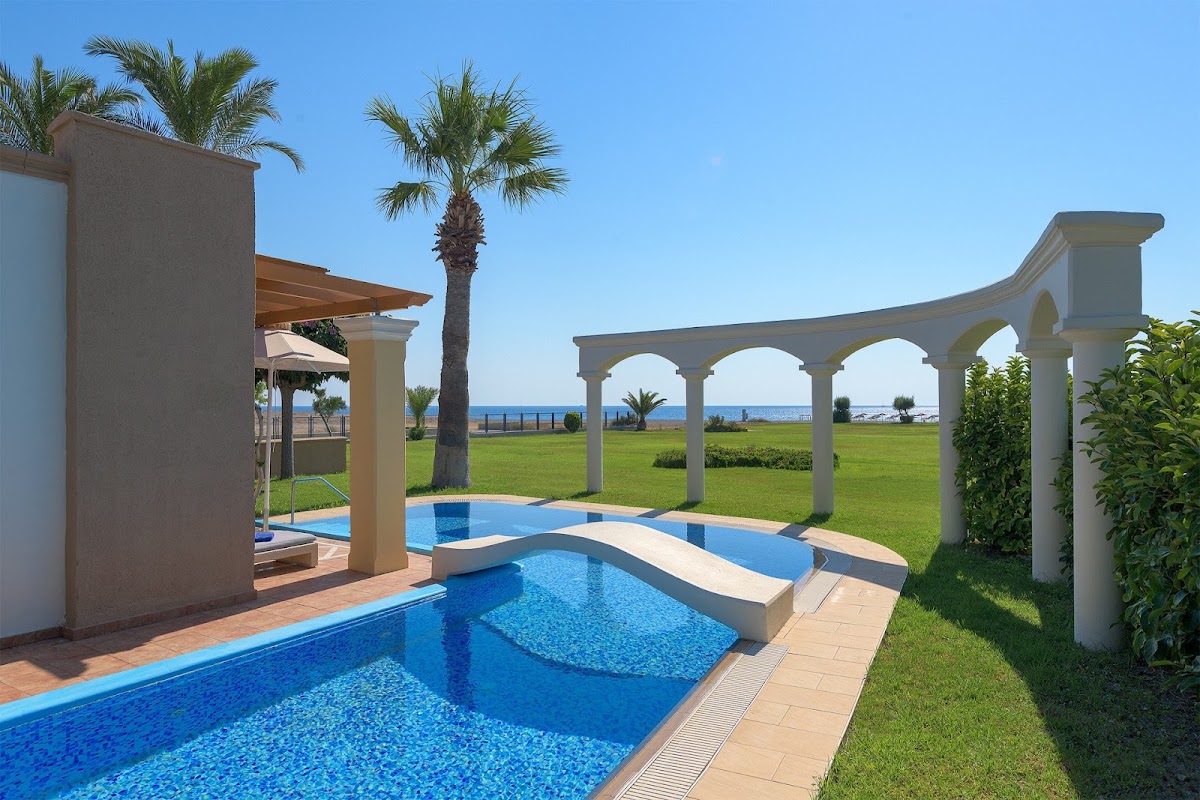 Multiple swimming pools with white columns and shade structures overlook manicured lawns and the Mediterranean Sea at Atrium Palace Thalasso Spa Resort.