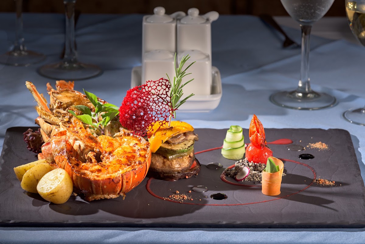 Fine dining plated entrée featuring roasted lobster, red fruit garnish, and herb accents on dark slate with wine glasses and white napkins.