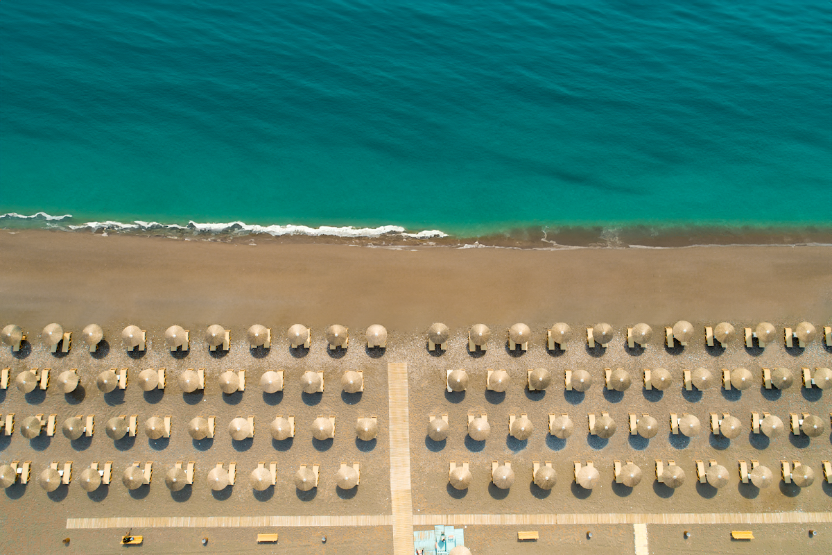 Overhead view of Atrium Palace Thalasso Spa Resort & Villas beach with rows of cream umbrellas and loungers on sand facing turquoise water and white-capped waves.