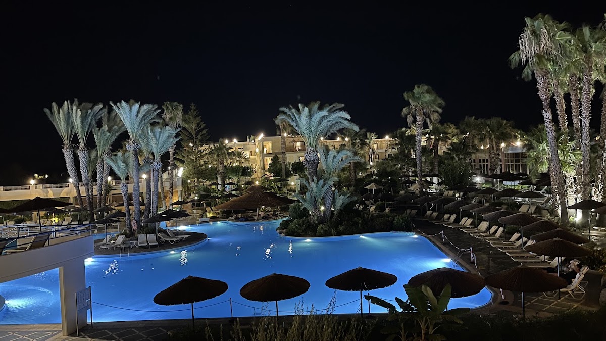 Illuminated resort pool at night surrounded by palm trees, loungers, and black umbrellas with lit buildings in the background.