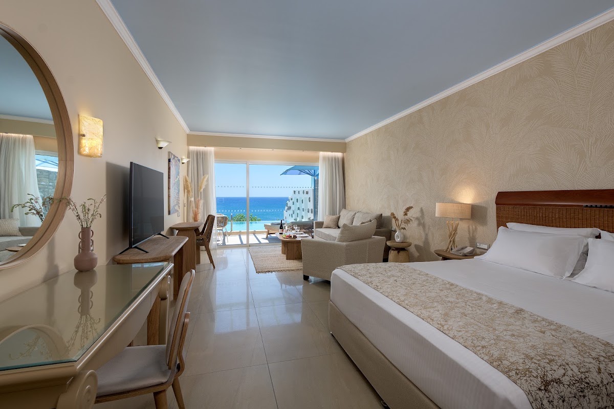 Resort guest room with queen bed, seating area, and ocean view balcony overlooking the Mediterranean Sea.